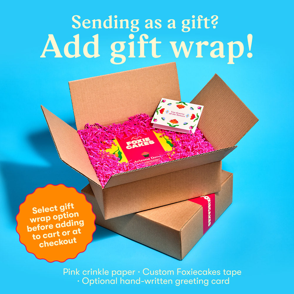 Open cardboard box with pink crinkle paper and a custom Foxiecakes tape and a floral greeting card on a blue background, with text "Sending as a gift? Add gift wrap! Select gift wrap option before adding to cart or at check out."