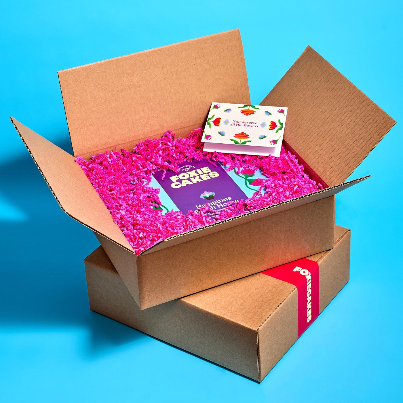 Open box with pink crinkle paper paper and a card and pink logo tape on a blue background, with text promoting gifting service.