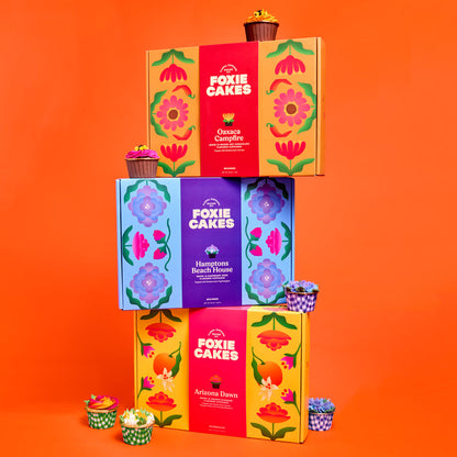 Stack of colorful 'Foxiecakes' floral cupcake kit boxes with floral designs and cupcakes on an orange background
