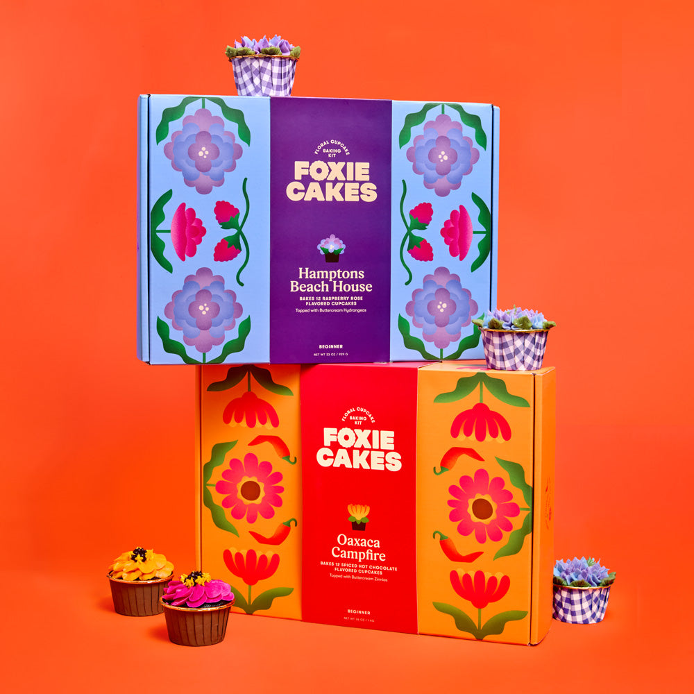 Stack of colorful 'Foxiecakes' floral cupcake kit boxes with floral designs and cupcakes on an orange background