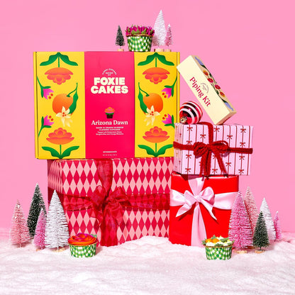 Decorative Foxiecakes Arizona Dawn baking kit gift box with festive pink and red wrapped Christmas packages, miniature trees, snow, ornaments and other winter holiday elements on a pink background.