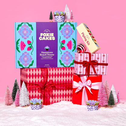 Decorative Foxiecakes Hamptons Beach House baking kit gift box with festive pink and red wrapped Christmas packages, miniature trees, snow, ornaments and other winter holiday elements on a pink background.