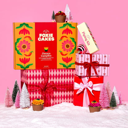 Decorative Foxiecakes Oaxaca Campfire baking kit gift box with festive pink and red wrapped Christmas packages, miniature trees, snow, ornaments and other winter holiday elements on a pink background.