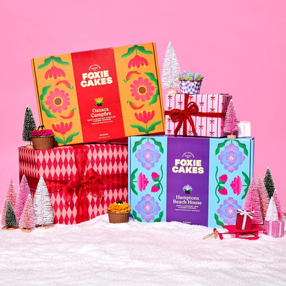 Two Foxiecakes baking kit boxes from the Starter Duo bundle with festive pink and red wrapped Christmas packages, miniature trees, snow, ornaments and other winter holiday elements on a pink background.