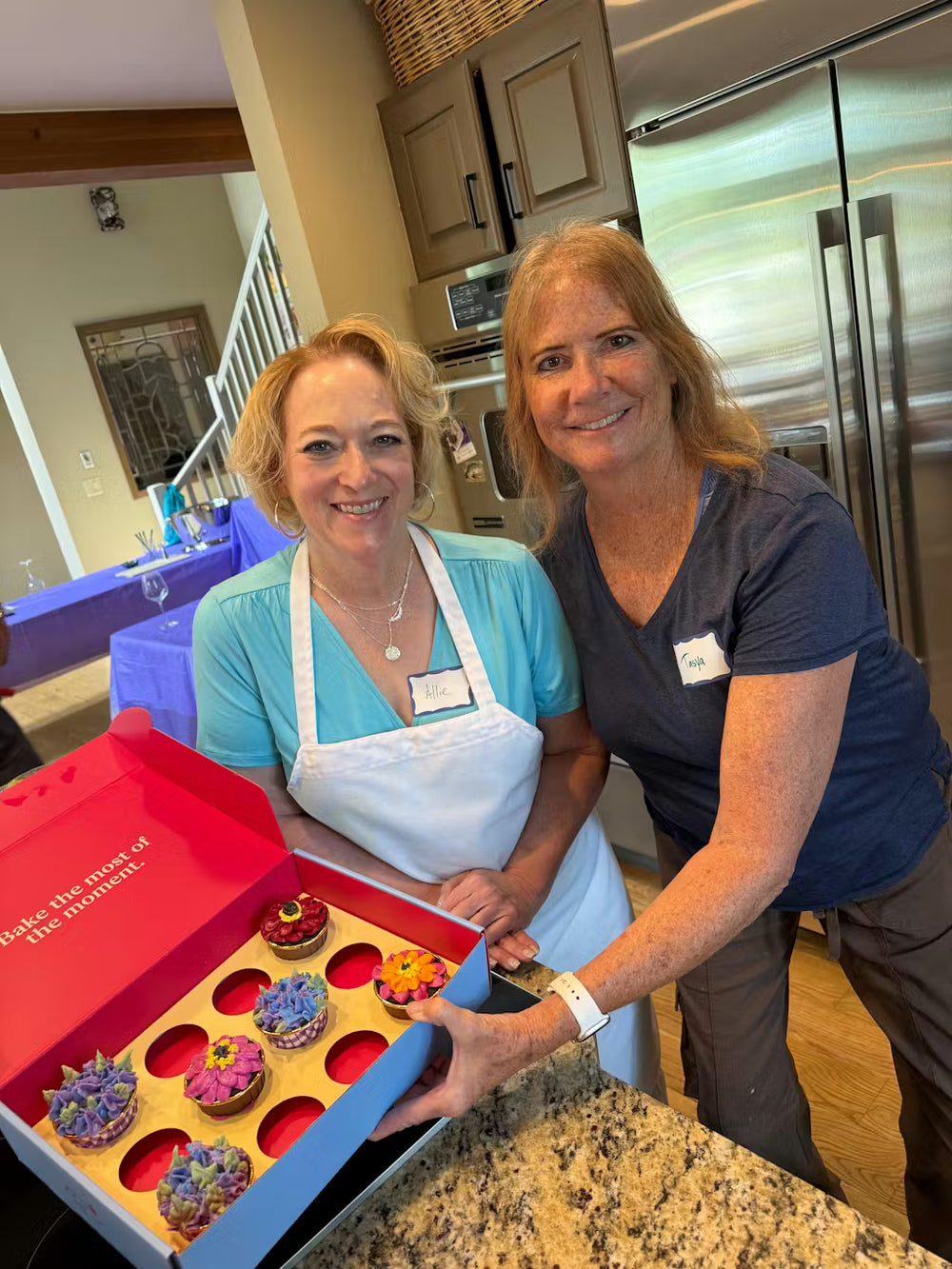 Two women in a kitchen show off a box of floral cupcakes they made