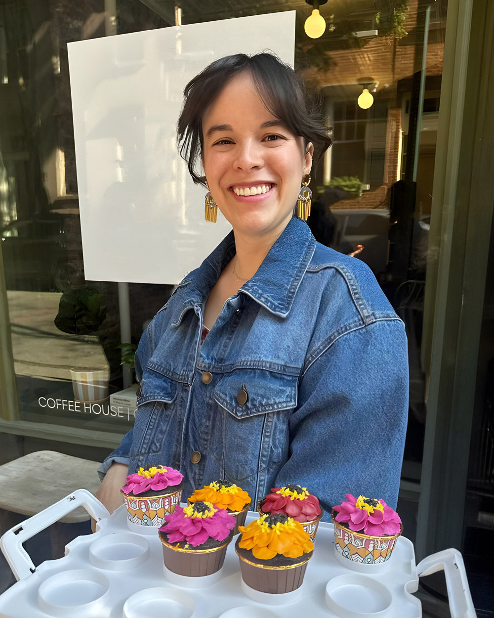 Photo of Foxiecakes founder Biz Jones in a denim jacket standing behind a display of cupcakes in front of a coffee shop.