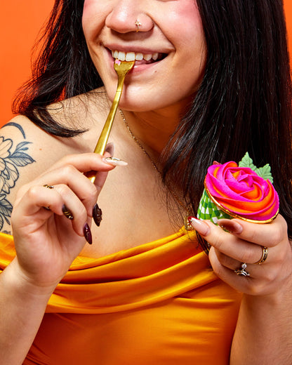 Woman eating a colorful buttercream rose cupcake with a gold fork against an orange background