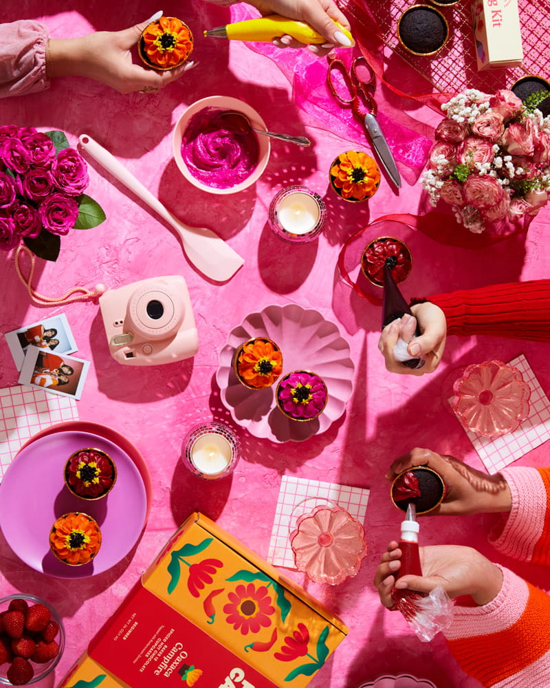 Colorful table setting with zinnia flower cupcakes, flowers, candles, and decorative items on a pink surface. Hands reach in to pipe buttercream flowers onto cupcakes