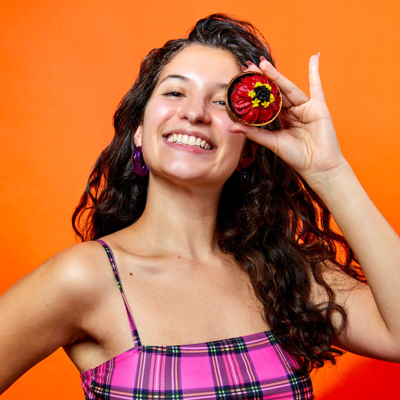 Woman holding a decorative buttercream flower zinnia cupcake near her eye against an orange background