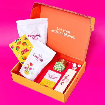 Foxiecakes Arizona Dawn floral cupcake kit open box with cake mix, frosting mix, orange blossom water, food coloring, and piping tools on a pink  background