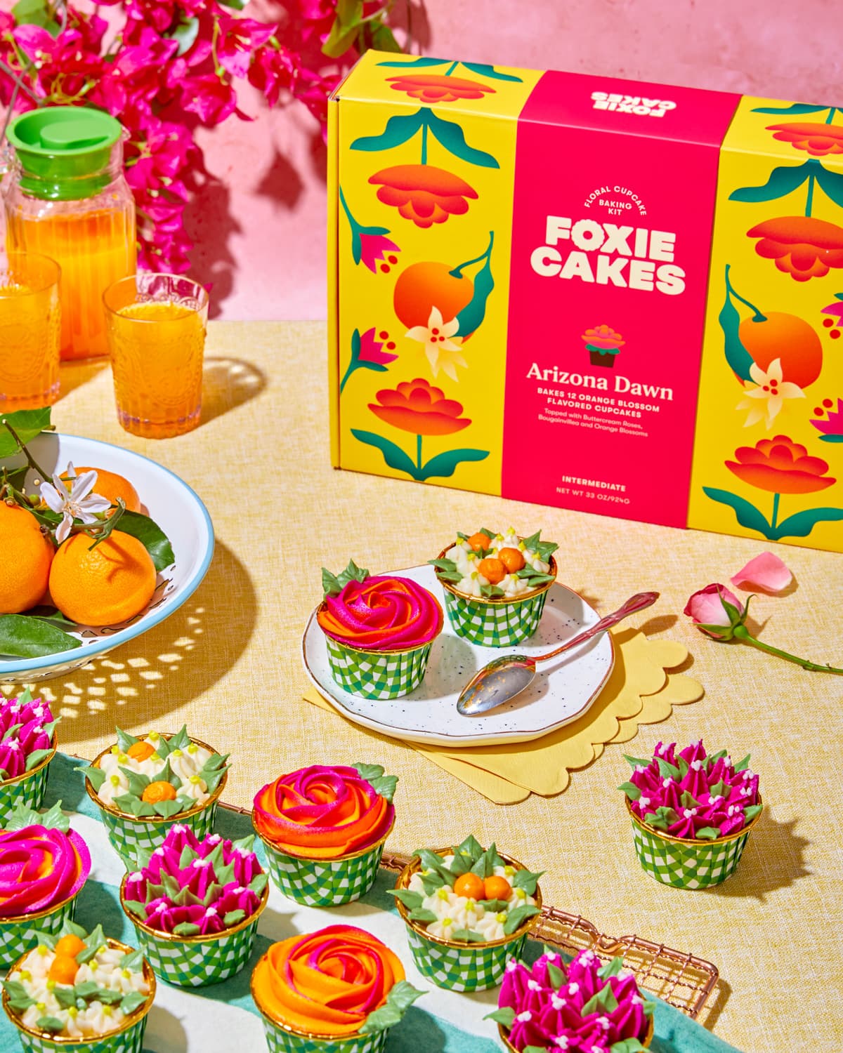Colorful cupcakes and 'Foxiecakes' Arizona Dawn floral cupcake kit packaging on a sunny table style with buttercream flower cupcakes, oranges and orange juice. On a yellow tablecloth with a pink plaster background.