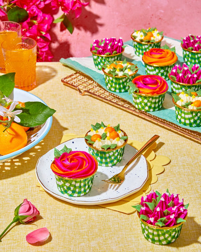 Colorful flower cupcakes on a sunny table style with buttercream flower cupcakes, oranges and orange juice. On a yellow tablecloth with a pink plaster background.