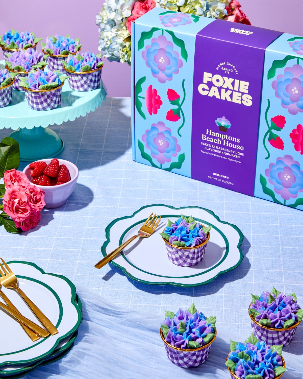 Raspberry rose cupcake with purple and blue hydrangea-style buttercream on a decorative plate, styled in natural outdoor lighting with hydrangea and rose florals, on a blue table cloth with Foxiecakes Hamptons Beach House packaging in a elegant garden-inspired setting. 