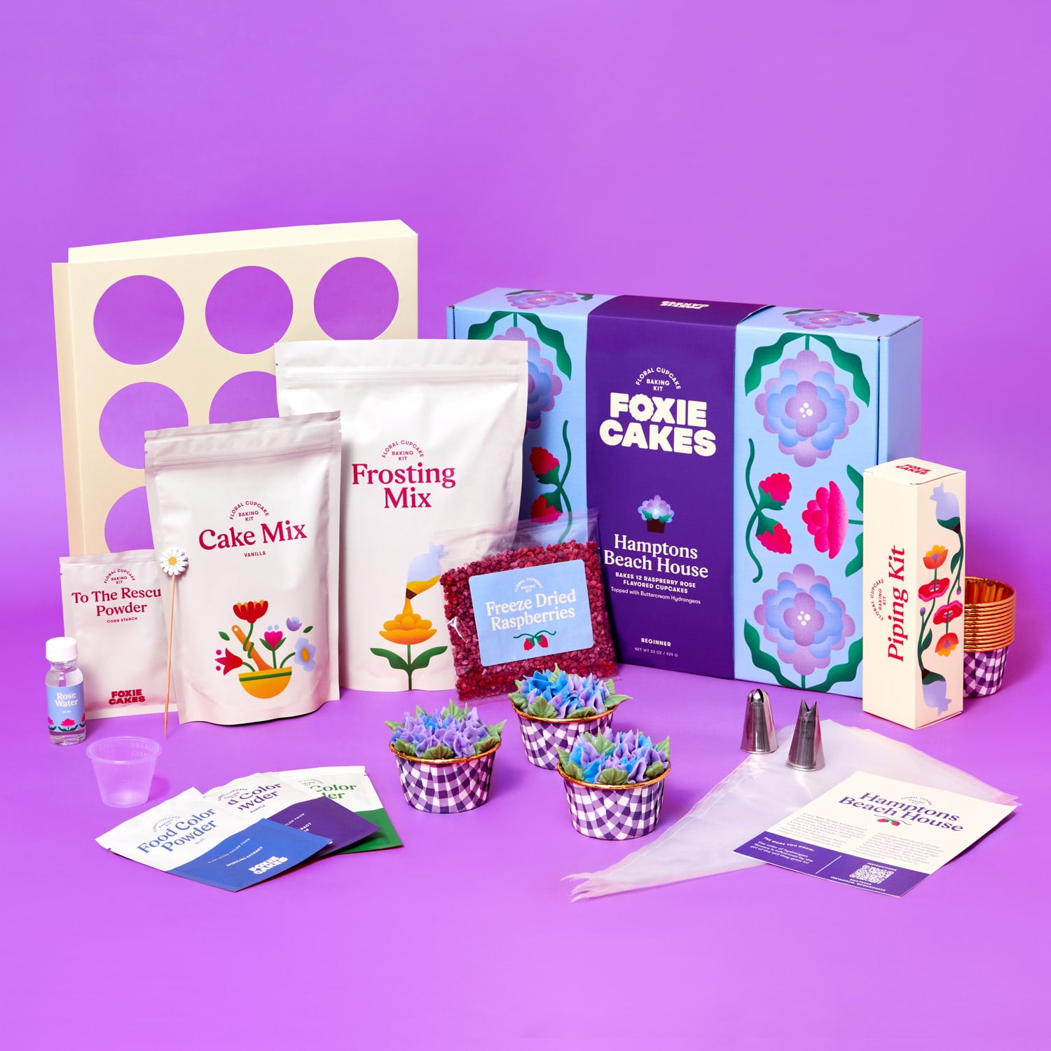 Foxiecakes Hamptons Beach House floral cupcake kit contents displayed, including cake mix, frosting mix, piping tips, food color powders, freeze-dried raspberries, baking tools, and decorated raspberry rose cupcakes topped with hydrangea-style buttercream next to closed kit, on a purple background. 