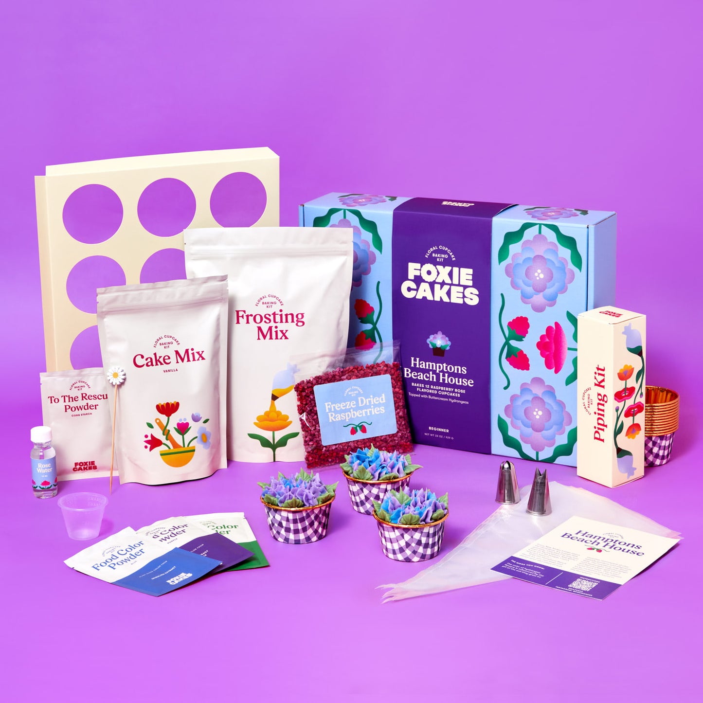 Foxiecakes Hamptons Beach House floral cupcake kit contents displayed, including cake mix, frosting mix, piping tips, food color powders, freeze-dried raspberries, baking tools, and decorated raspberry rose cupcakes topped with hydrangea-style buttercream next to closed kit, on a purple background. 