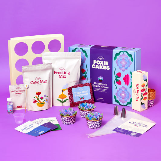Foxiecakes Hamptons Beach House floral cupcake kit contents displayed, including cake mix, frosting mix, piping tips, food color powders, freeze-dried raspberries, baking tools, and decorated raspberry rose cupcakes topped with hydrangea-style buttercream next to closed kit, on a purple background. 