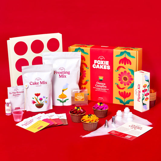 Foxiecakes Oaxaca Campfire floral cupcake kit contents displayed, including cake mix, frosting mix, piping tips, food color powders, spice mix, baking tools, and decorated spiced hot chocolate cupcakes topped with zinnia-style buttercream next to closed kit, on a red background. 