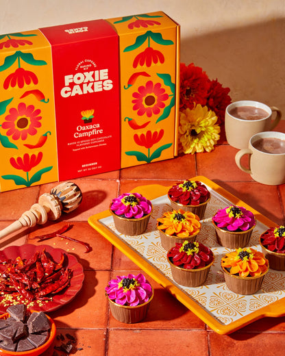 Foxiecakes Oaxaca Campfire floral cupcake kit packaging on a table  with spiced hot chocolate cupcakes with vibrant pink orange and red zinnia-style buttercream flowers on a decorative orange tile plate, styled in sunset golden hour lighting on orange tile, with mugs of hot chocolate and side plates of chiles and chopped chocolate.