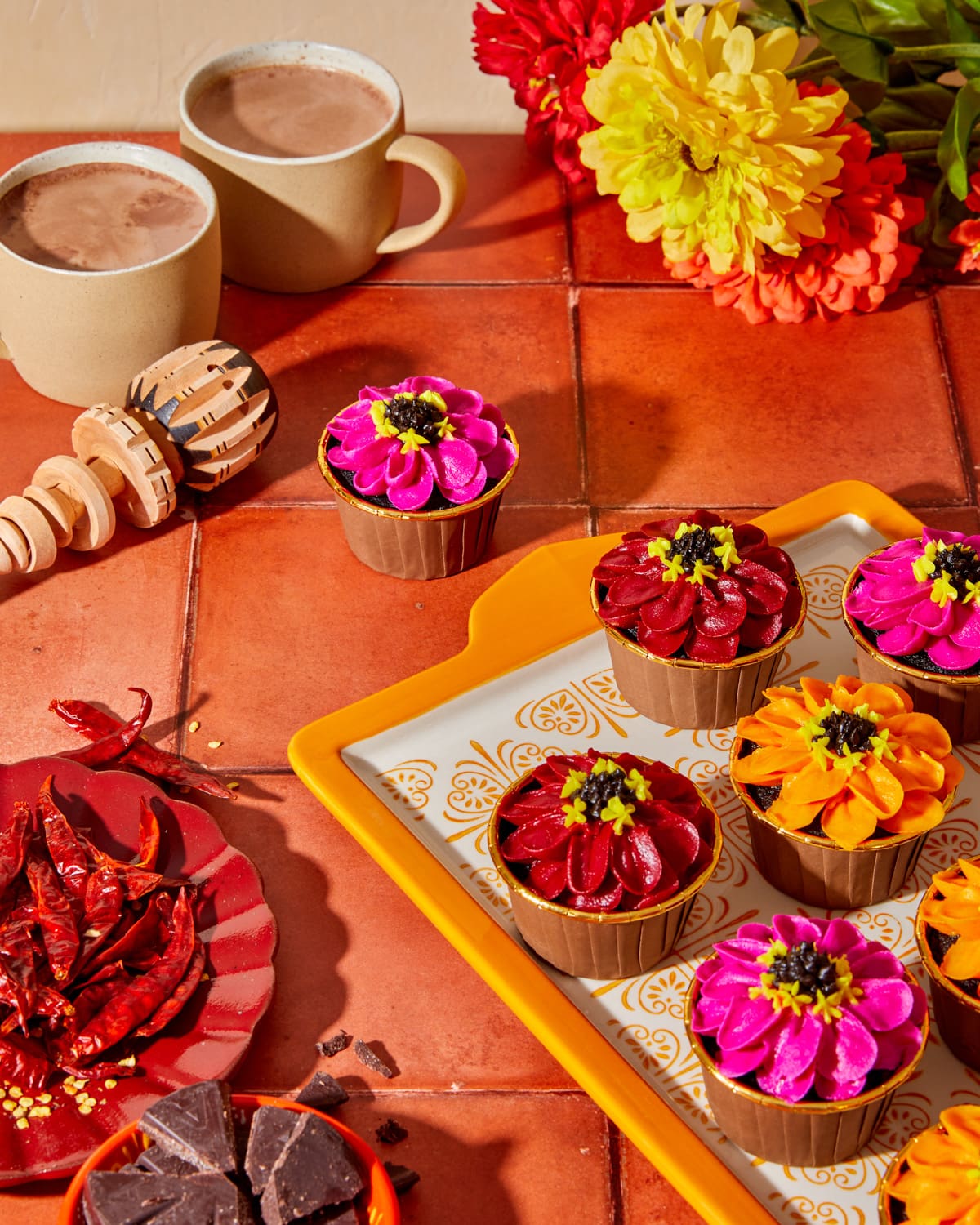 Spiced hot chocolate cupcakes with vibrant pink orange and red zinnia-style buttercream flowers on a decorative orange tile plate, styled in sunset golden hour lighting on orange tile, with mugs of hot chocolate and side plates of chiles and chopped chocolate.