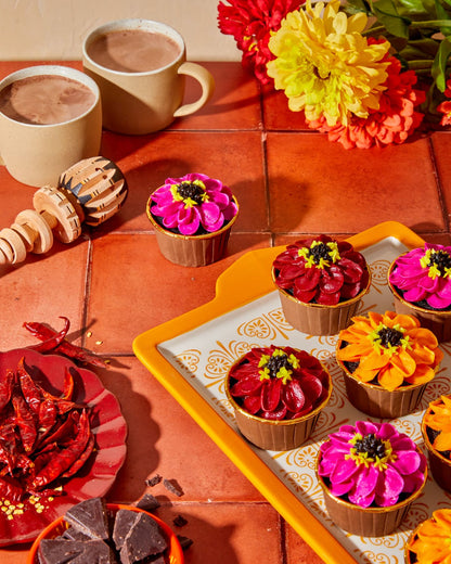 Spiced hot chocolate cupcakes with vibrant pink orange and red zinnia-style buttercream flowers on a decorative orange tile plate, styled in sunset golden hour lighting on orange tile, with mugs of hot chocolate and side plates of chiles and chopped chocolate.