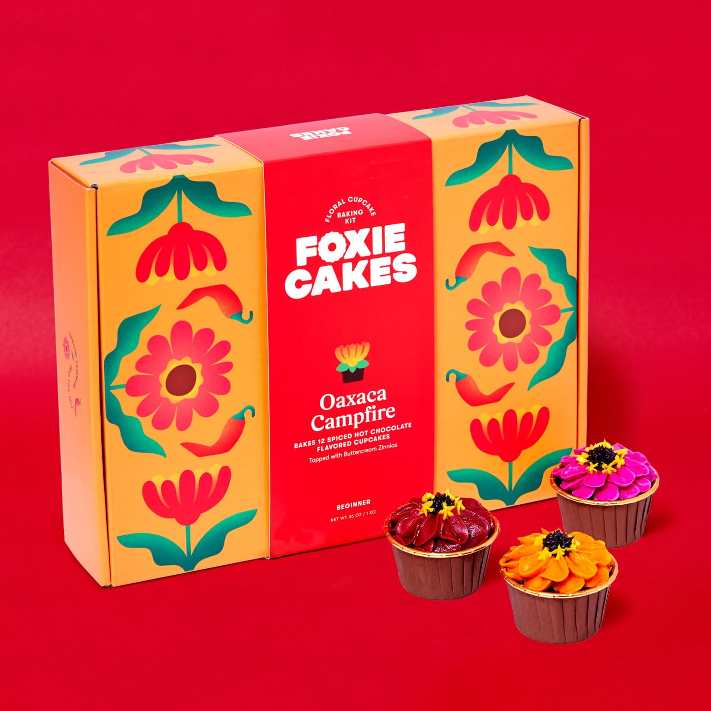 Foxiecakes Oaxaca campfire floral cupcake baking kit box displayed with three decorated spiced hot chocolate cupcakes featuring zinnia-style buttercream frosting, on a red background. Beginner-friendly DIY cupcake decorating kit for a creative at-home baking experience