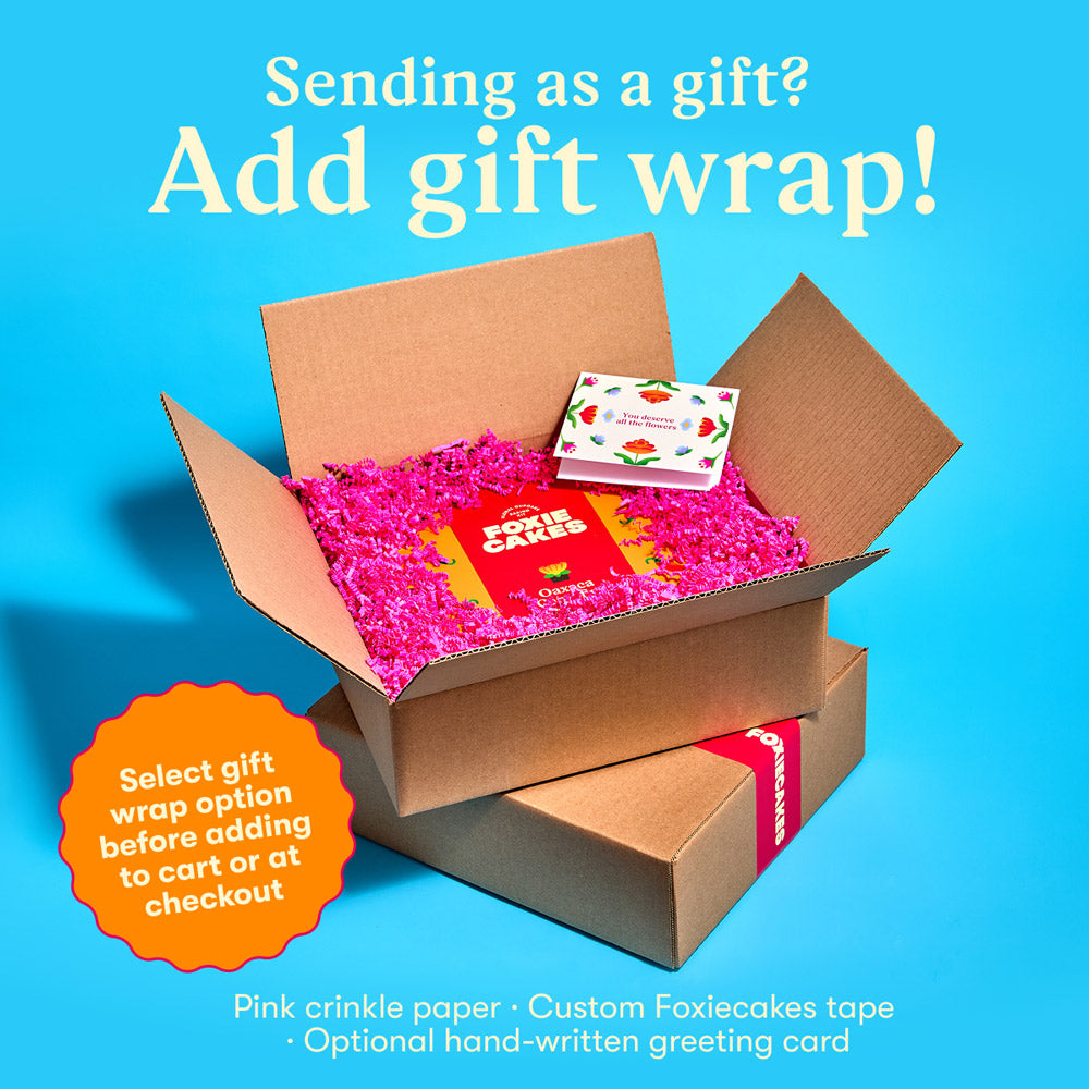 Open cardboard box with pink crinkle paper and a custom Foxiecakes tape and a  floral greeting card on a blue background, with text "Sending as a gift? Add gift wrap! Select gift wrap option before adding to cart or at check out."