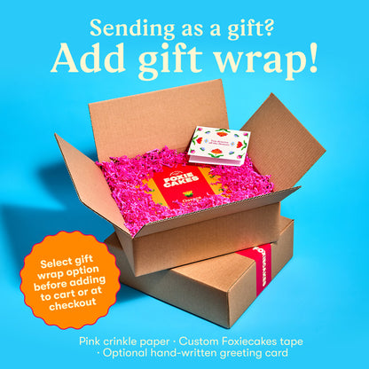Open cardboard box with pink crinkle paper and a custom Foxiecakes tape and a  floral greeting card on a blue background, with text "Sending as a gift? Add gift wrap! Select gift wrap option before adding to cart or at check out."