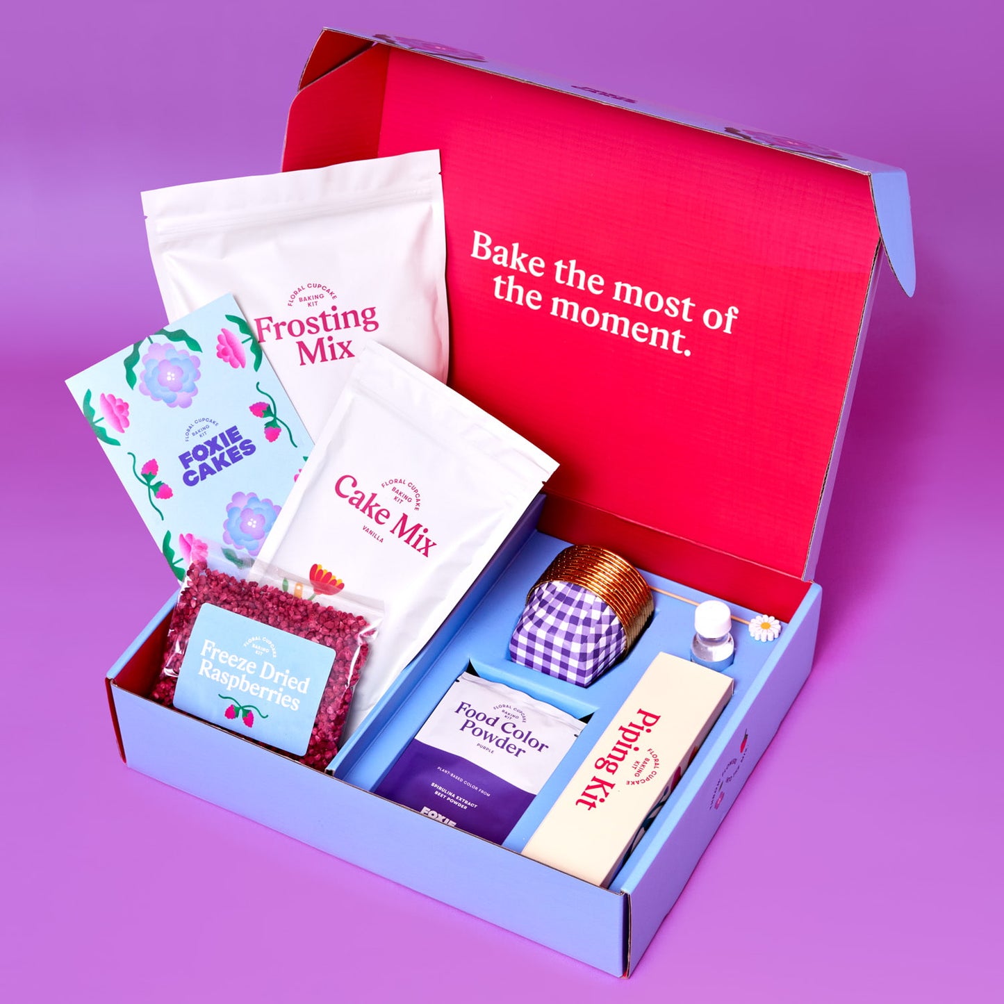 Foxiecakes Hamptons Beach House floral cupcake kit open box with cake mix, frosting mix, freeze-dried raspberries, rose water, food coloring, and piping tools on a purple  background